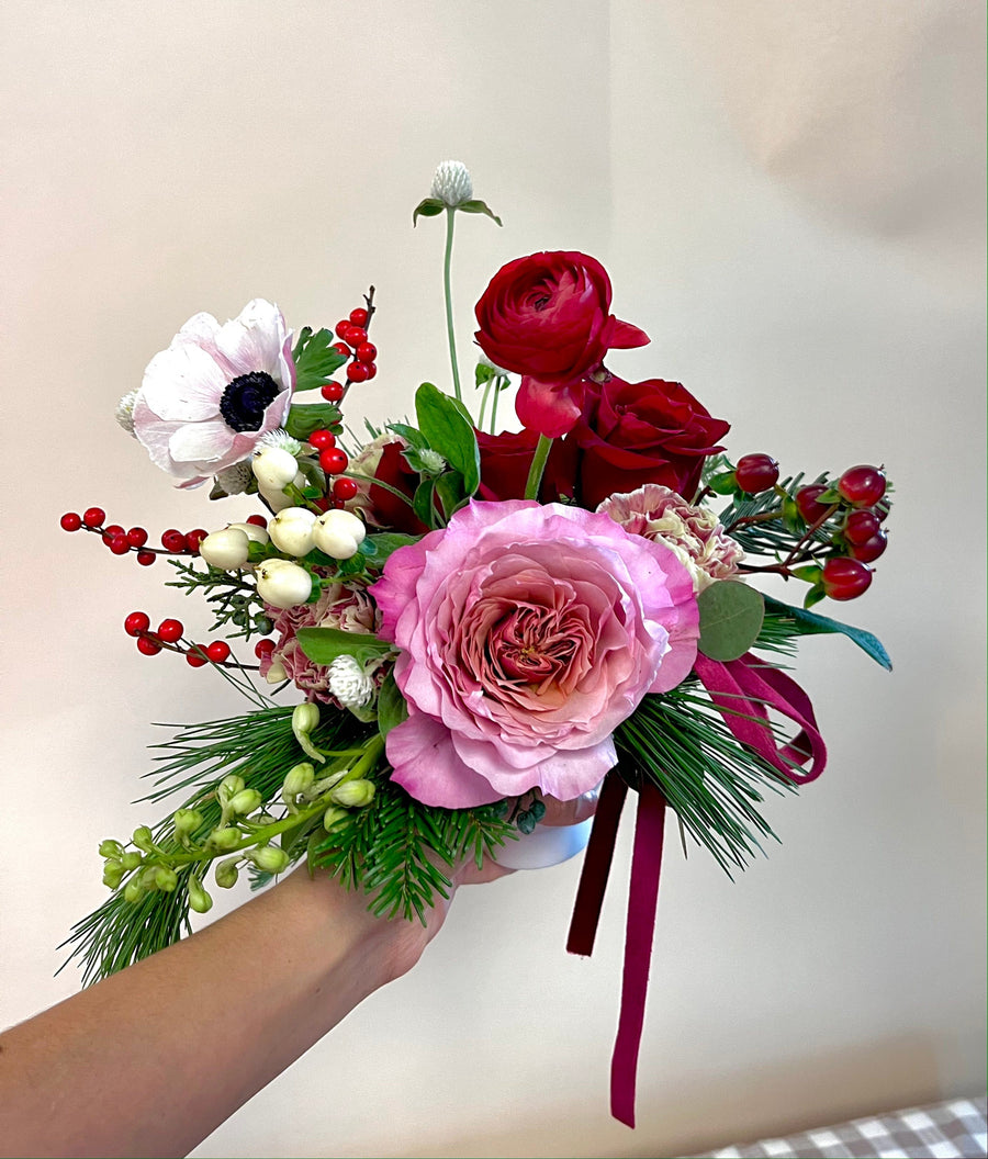 In a small white compote with mauve garden rose, red ranunculus, 'freedom' red roses, red spray roses, white anemone, white hypericum berries, ilex berries, variegated carnation, pine greenery, cedar greenery. Holiday season greeting gift here in Houston.