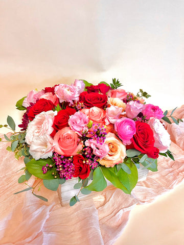 A floral arrangement featuring red roses, coral roses, peach roses, pink ranunculus, hot pink ranunculus, pink lisianthus, waxflower in a wooden box.