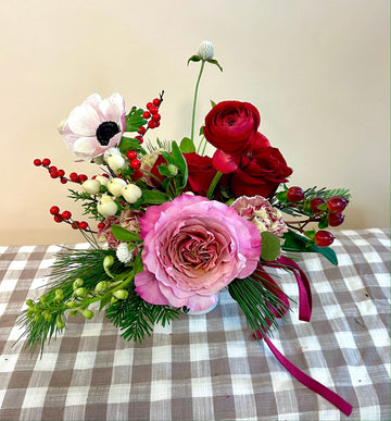 A floral arrangement featuring a variety of flowers including pink and red roses, white anemone, and greenery, presented in a small white compete.