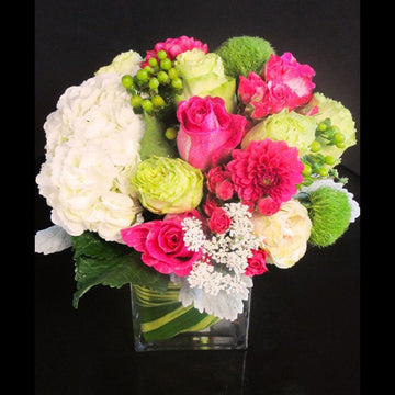 White hydrangea, roses in green and hot pink with green hypericum berries in a clear cube vase. 