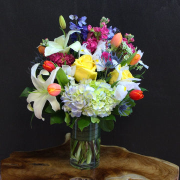 Yellow roses, white lilies, white irises and more in a cylinder vase bring joy to someone’s special day or birthday.