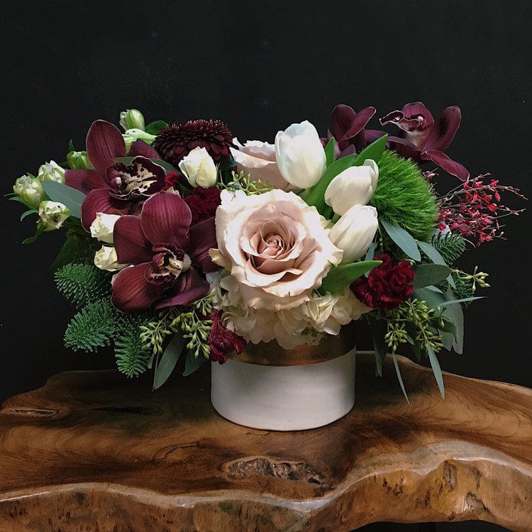 Holiday arrangement with 'Quicksand' roses, white hydrangeas, green dianthus, burgundy cymbidium orquids, white tulips, white alstromerias, ilex, spray roses, celosias, burgundy pompon gerberas, seeded eucalyptus, and seasonal greeneries in a white and gold ceramic vase. Beautiful holiday arrangement for any birthday, anniversary, or just because. Available for same day delivery and pickup in Houston Heights 