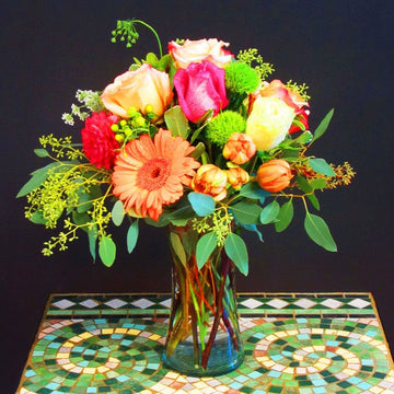 Hot pink roses, orange dahlias, orange tulips and gerberas fresh eucalyptus in a concave glass vase 