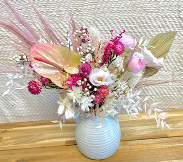 A dried palm leaf, mauve pampas grass, vibrant hot pink dried strawflower, pink lisianthus, pink ranunculus, dried tallow berries, and preserved ruscus in a white ceramic vase.