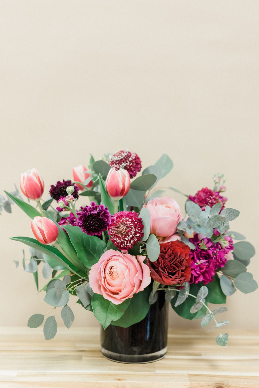 Valentine arrangment with garden roses, scabiosa pods, tulips, stock, and eucalyptus greenery, elegantly arranged in a black cylinder vase.